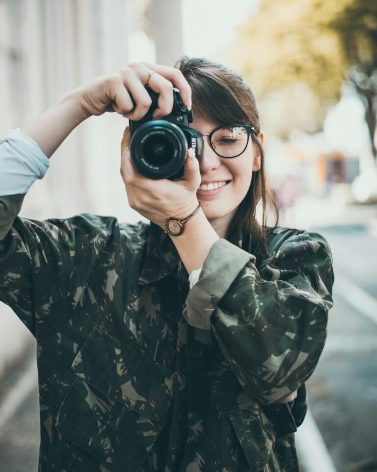 A smiling woman in glasses takes a photo outdoors, capturing a moment.